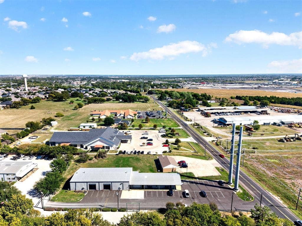 911 W Holiday Drive, Fate, TX 75132 Aerial perspective of the property featuring a metal-sided commercial warehouse, an asphalt parking lot, two prominent steel support pillars, and direct frontage to a paved roadway