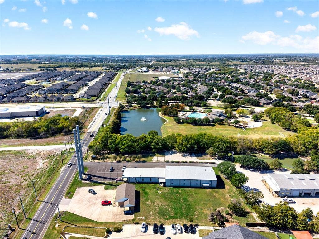 911 W Holiday Drive, Fate, TX 75132 Aerial perspective of property featuring paved surface parking, metal-roof structure, and expansive open green space