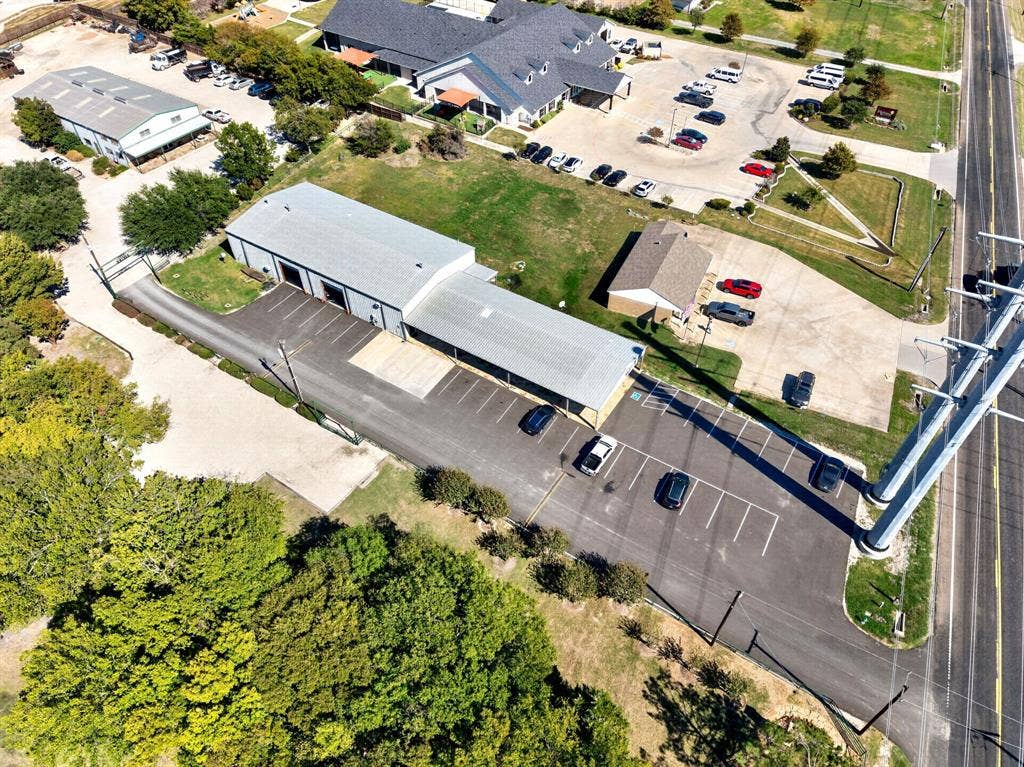911 W Holiday Drive, Fate, TX 75132 Commercial property featuring a metal-sided building, expansive paved parking lot, covered exterior loading area, direct road access, and mature perimeter trees