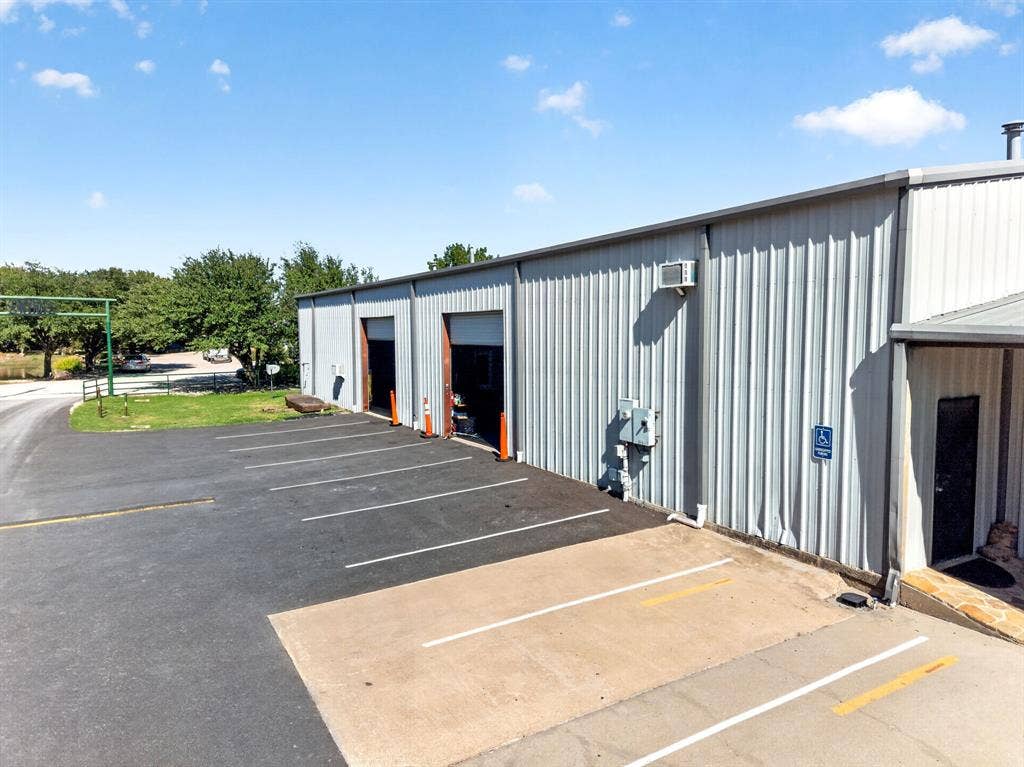 911 W Holiday Drive, Fate, TX 75132 Metal-paneled exterior building featuring two wide bay entrances, paved parking area with designated spaces, and established exterior lighting