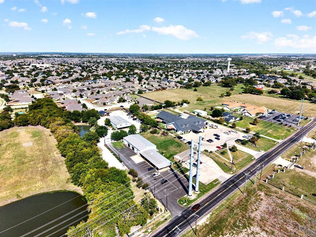 911 W Holiday Drive, Fate, TX 75132 Aerial perspective featuring multiple industrial-style metal structures, paved parking areas, covered canopy parking, and a natural pond situated on a large lot