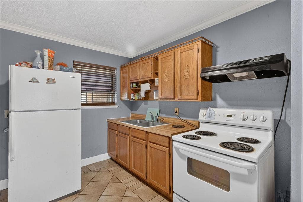 Kitchen featuring natural wood cabinetry, dual-basin stainless steel sink, tile flooring, and wall-mounted range hood