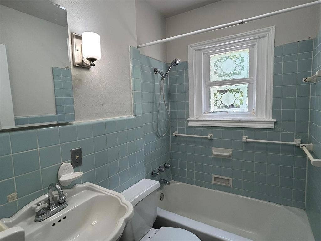 Bathroom featuring blue tile wall surround, integrated bathtub and shower combination, wall-mounted light fixture, pedestal sink, and privacy window with decorative glass detailing