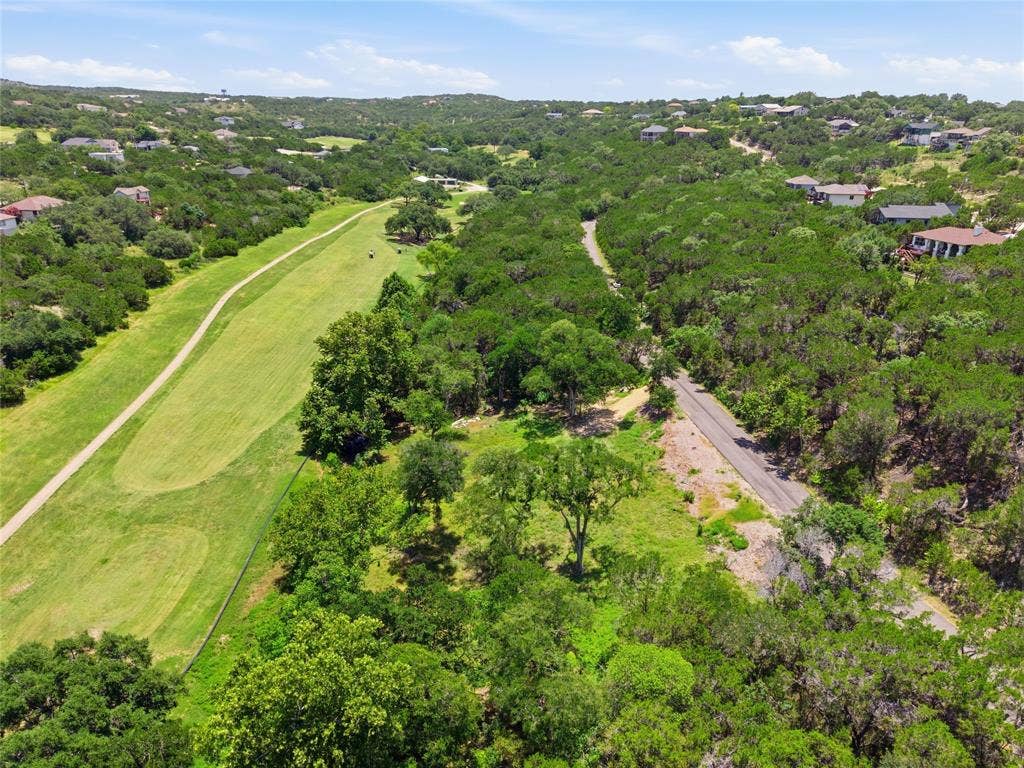 Aerial perspective of a sloping lot featuring mature trees, native brush, paved road access, and proximity to maintained green space