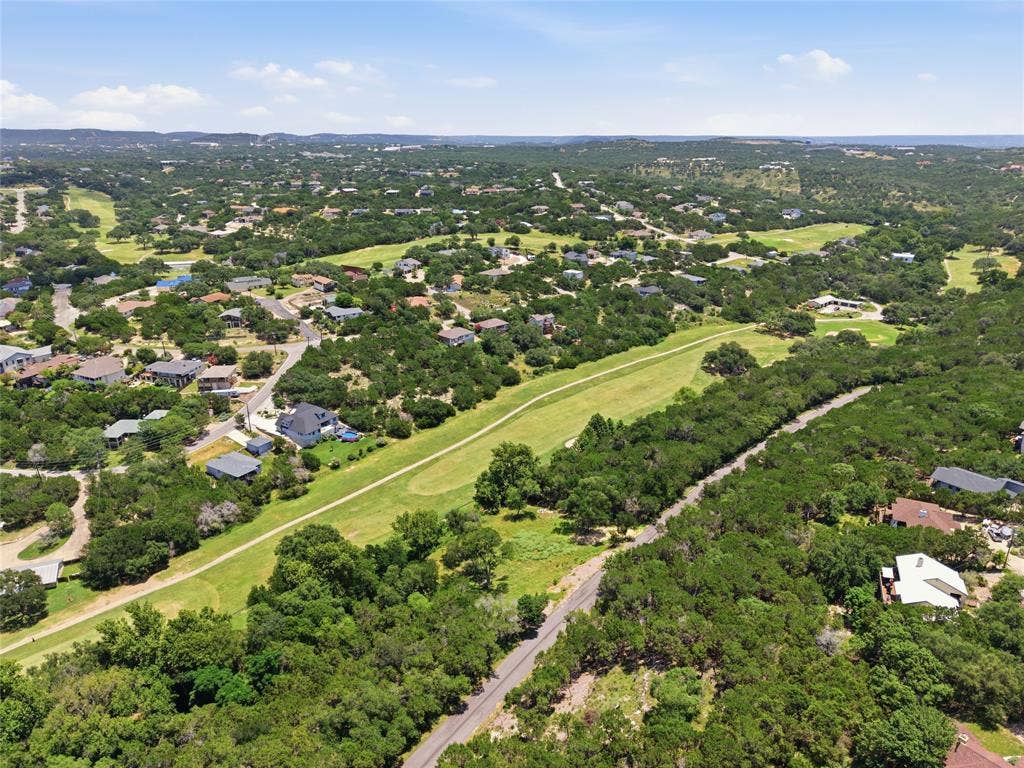 Aerial perspective of a sprawling landscape featuring expansive green terrain, dense clusters of mature trees, winding paved roads, and residential properties