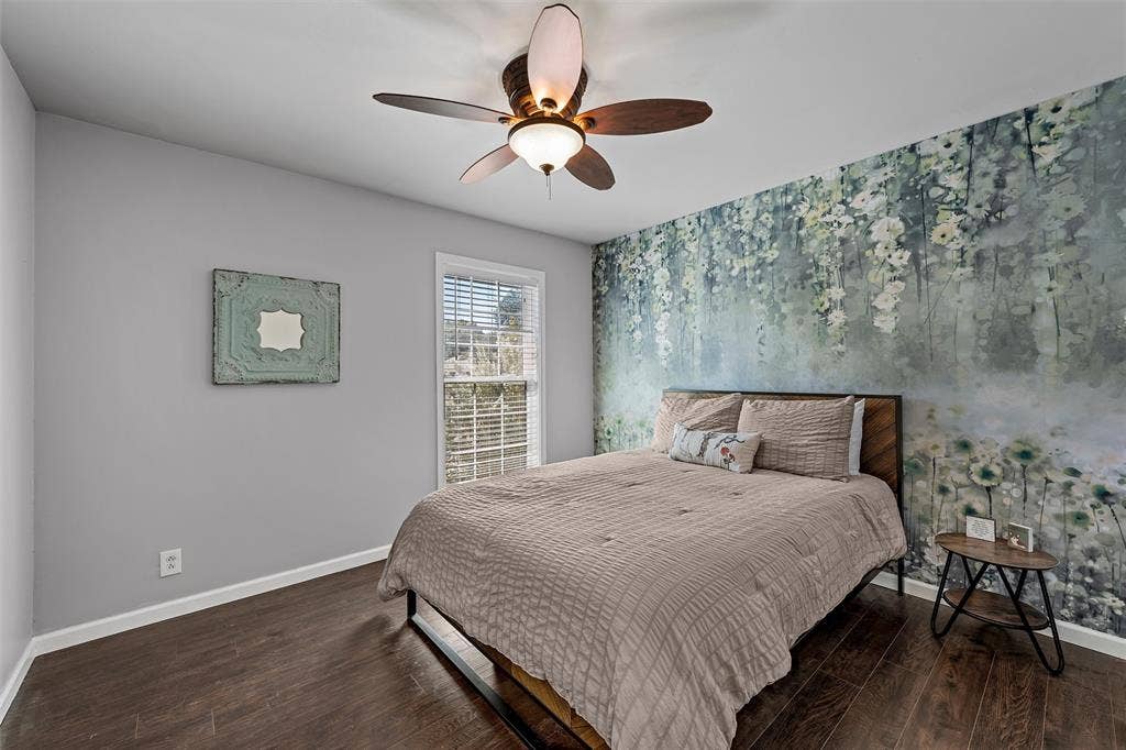 Room featuring wood-finish flooring, a multi-blade ceiling fan with dual-light fixtures, a single-hung window with horizontal blinds, and a botanical-patterned accent wall