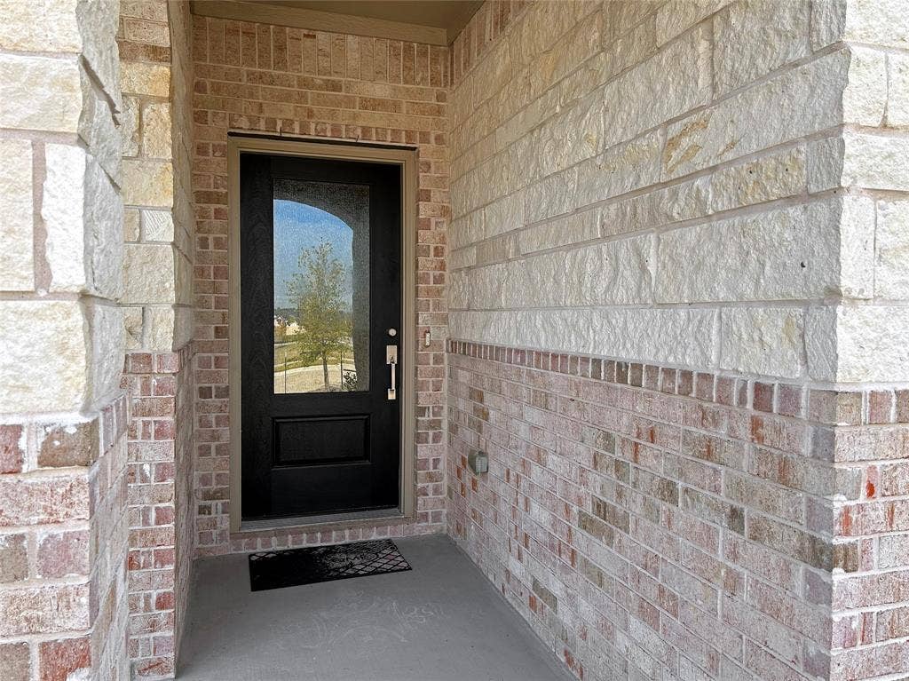 2504 Clyde Road, Celina, TX 75009 Covered entryway featuring mixed-masonry stone and brick facade, decorative solid-core front door with an integrated glass panel, and brushed metal hardware