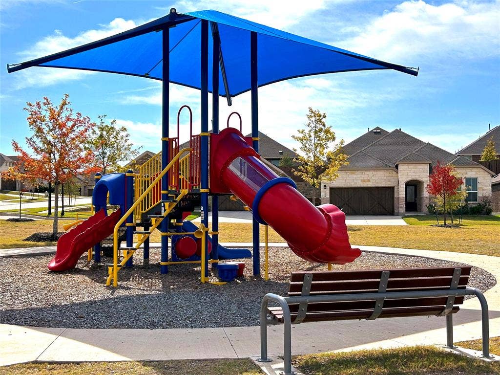 2504 Clyde Road, Celina, TX 75009 Front of house with a stone exterior as seen from the community recreational area. The recreation area features a structured play system with a blue shade canopy, integrated slides, a gravel base, a fixed metal and wood bench and interconnected sidewalks.