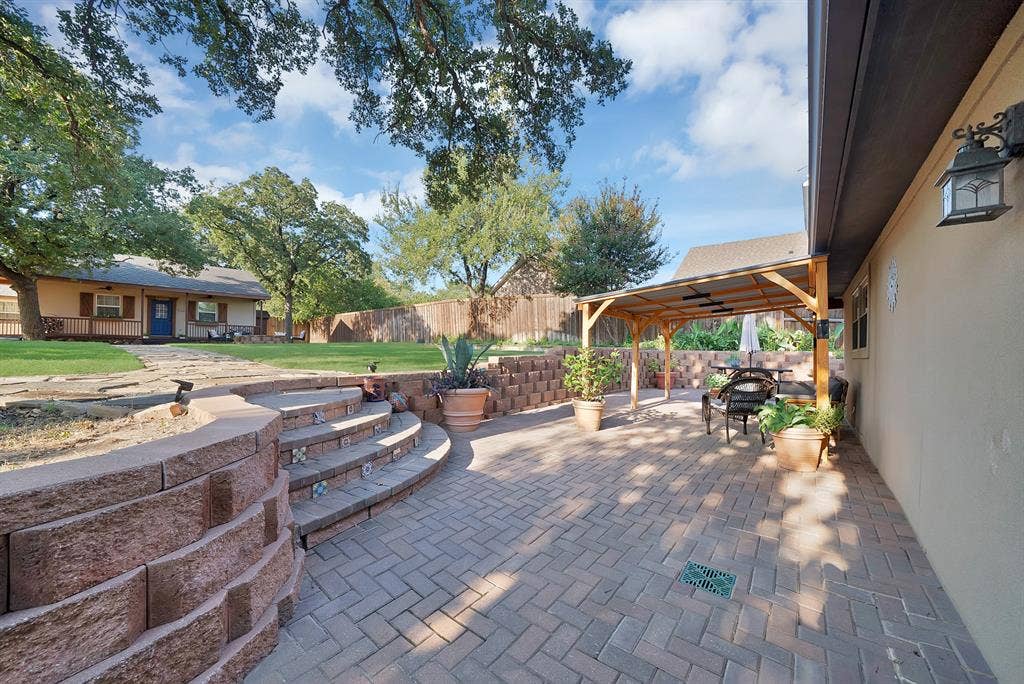 306 E Rochelle Boulevard, Irving, TX 75062 Herringbone-patterned paver patio featuring a timber-framed pergola, segmental retaining walls, decorative tiled steps, and a sodded lawn
