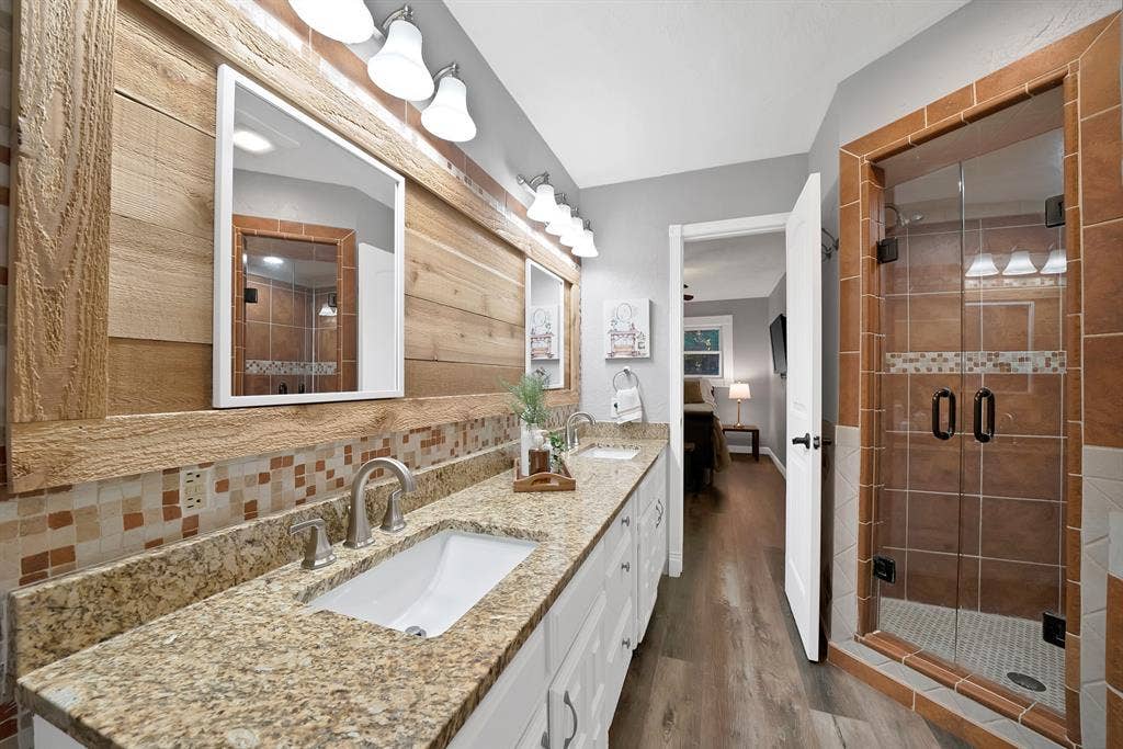 306 E Rochelle Boulevard, Irving, TX 75062 Dual-sink vanity with stone countertop, wood-plank wall accent, glass-enclosed shower with tiled surround, and wood-finish flooring