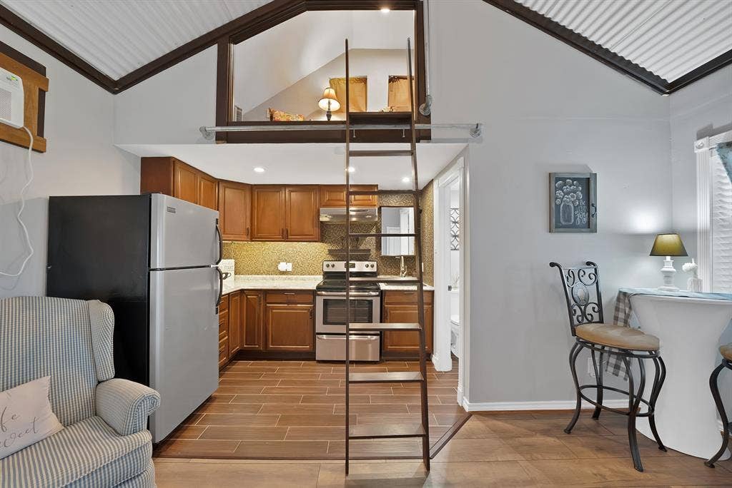 306 E Rochelle Boulevard, Irving, TX 75062 Kitchen featuring wood cabinetry, stainless steel appliances, tile backsplash, and wood-finish flooring, complemented by a structural ladder providing access to an elevated mezzanine level under a vaulted ceiling