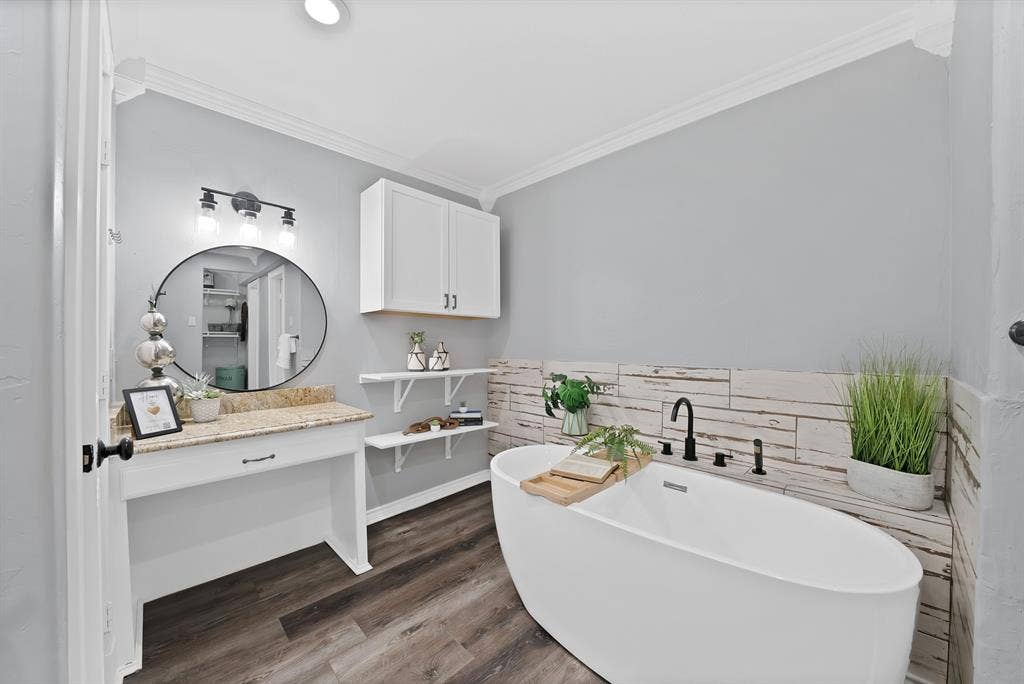306 E Rochelle Boulevard, Irving, TX 75062 Freestanding soaking tub featuring a matte black deck-mounted faucet, wood-finish flooring, white vanity with natural stone countertop, circular wall mirror, and crown molding