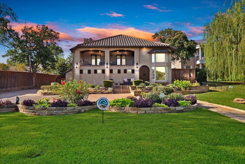 306 E Rochelle Boulevard, Irving, TX 75062 Two-story residence featuring a tile roof, an upper-level covered balcony, a bay window, and tiered stone landscaping with integrated garden beds