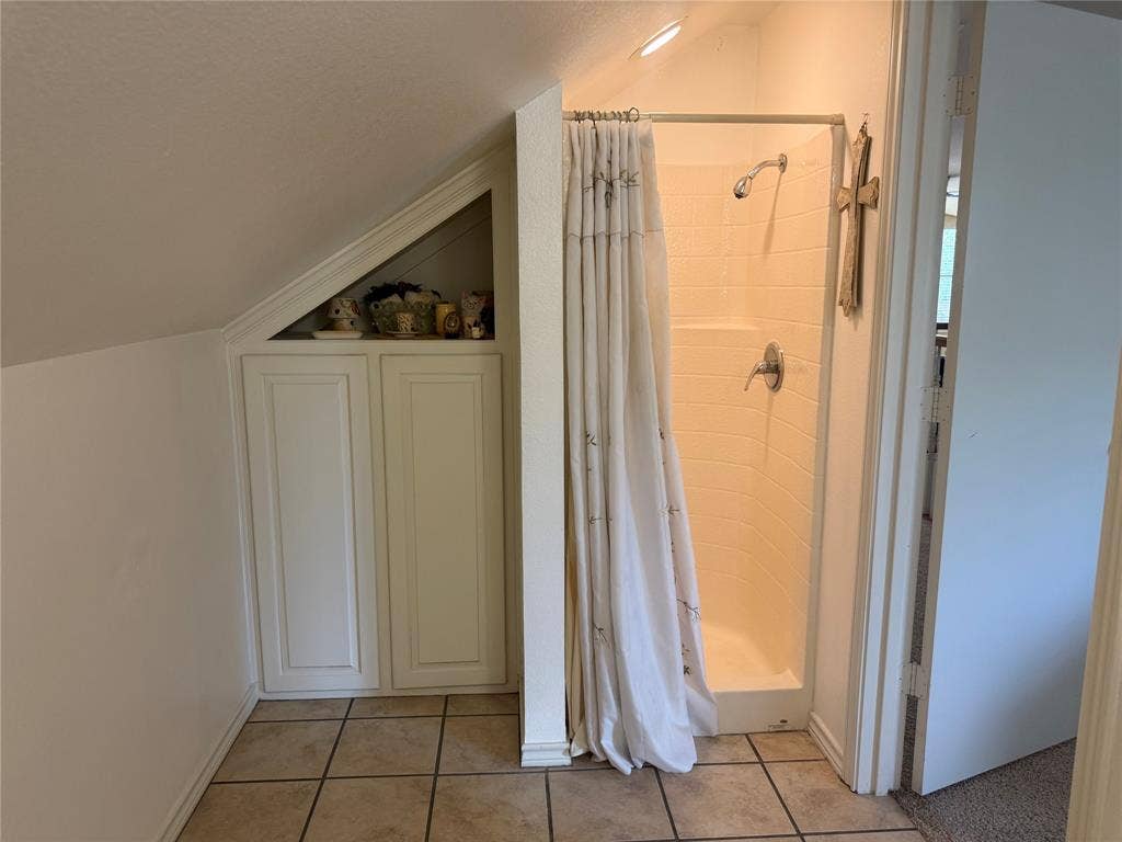 Shower stall featuring molded surround and metal fixtures, accompanied by built-in under-eave cabinetry with recessed shelving and tile flooring