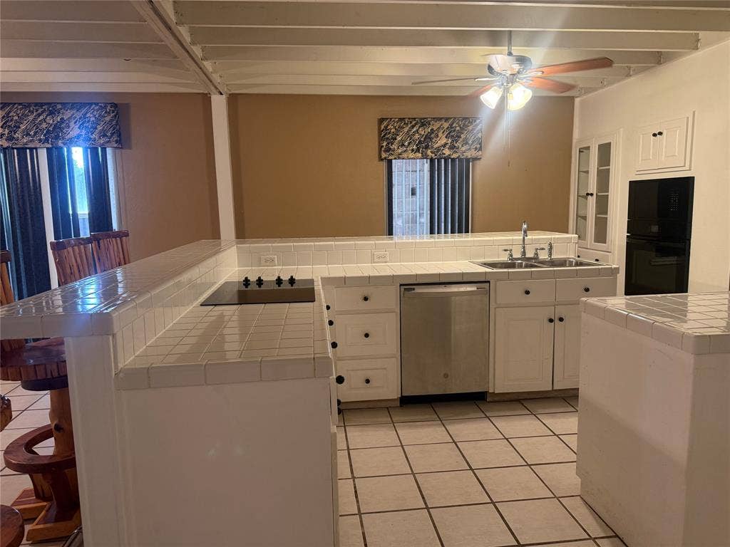 Kitchen featuring a breakfast bar, tile countertops, white cabinetry, stainless steel dishwasher, and built-in double oven