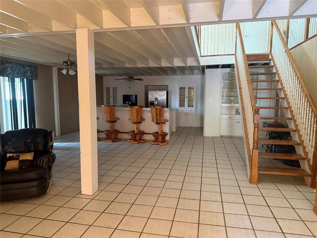 Open-concept interior featuring exposed beam ceilings, ceramic tile flooring, a central kitchen island with built-in cabinetry, and a staircase.