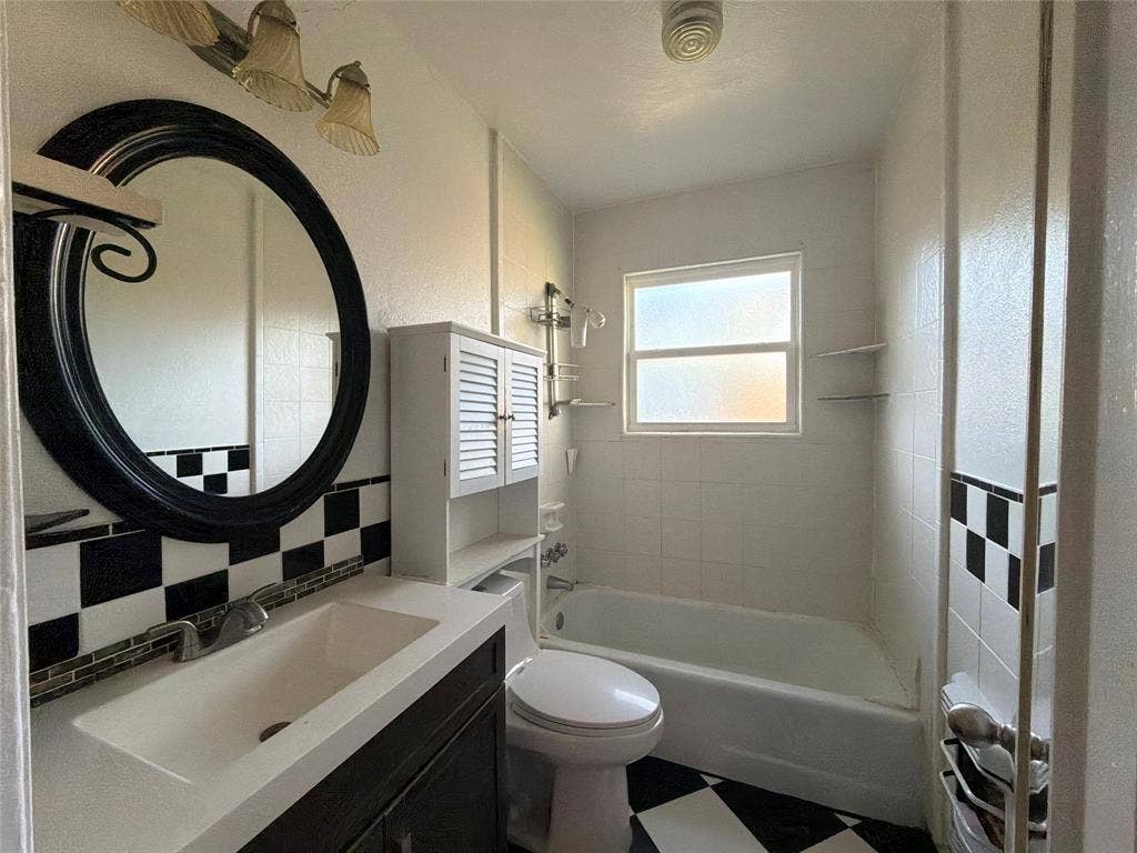 1819 Finley Road, Irving, TX 75062 Bathroom featuring a vanity with an integrated basin, a black-framed circular mirror, a built-in shuttered cabinet, a bathtub with a tiled surround, and checkered flooring