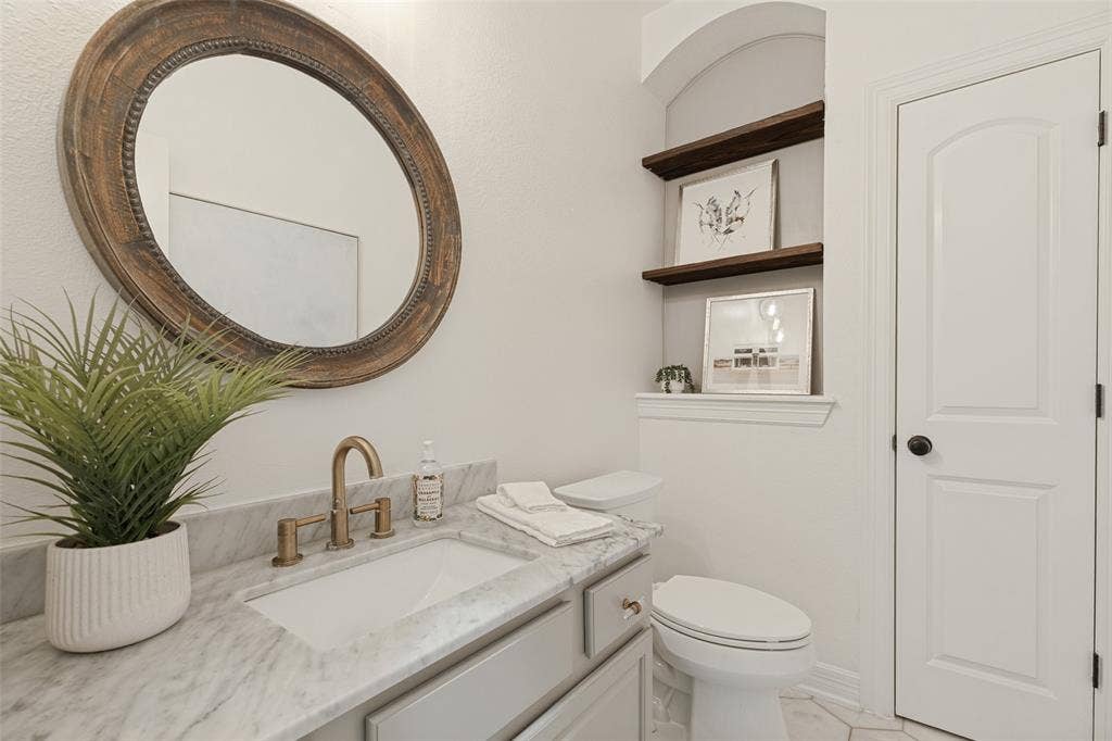 Vanity with stone countertop, undermount sink, and brass-finish widespread faucet, complemented by a circular wood-framed wall mirror, arched wall niche with wood shelving, and a paneled interior door linen closet