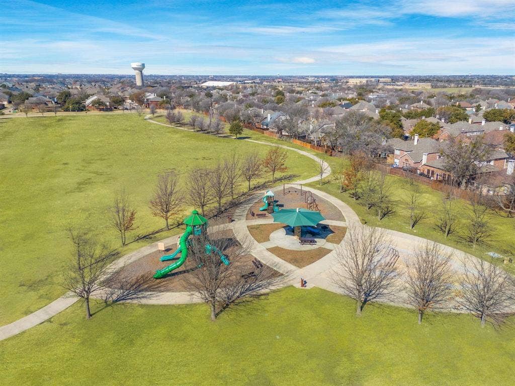 Public park area featuring a multi-feature playground structure, shaded pavilion, paved walking paths, and expansive green space