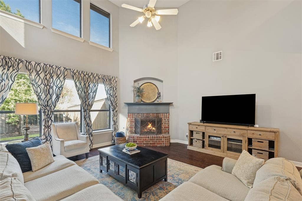 High-ceilinged living area featuring wood-finish flooring, floor-to-ceiling windows, a corner brick fireplace, and a ceiling fan