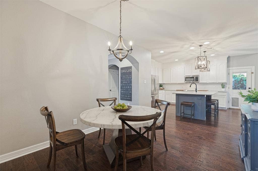 Open-concept area featuring wood-finish flooring, a kitchen island with stone countertop, white shaker-style cabinetry, arched architectural detailing, and dual pendant lighting fixtures