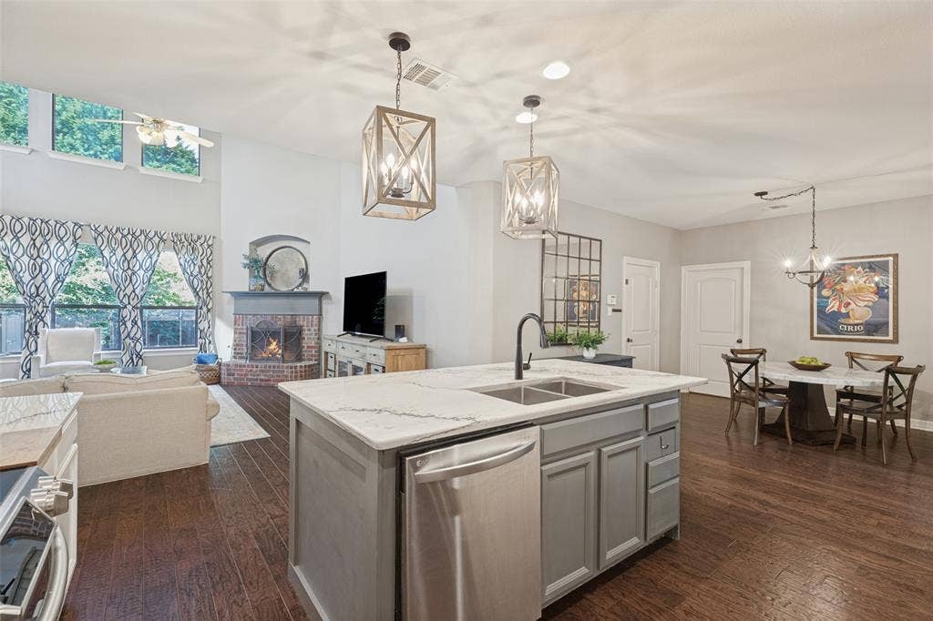 Kitchen island with a stone countertop, under-mount dual sink, stainless steel dishwasher, and two lantern-style pendant lights, featuring wood-finish flooring and a corner brick-surround fireplace