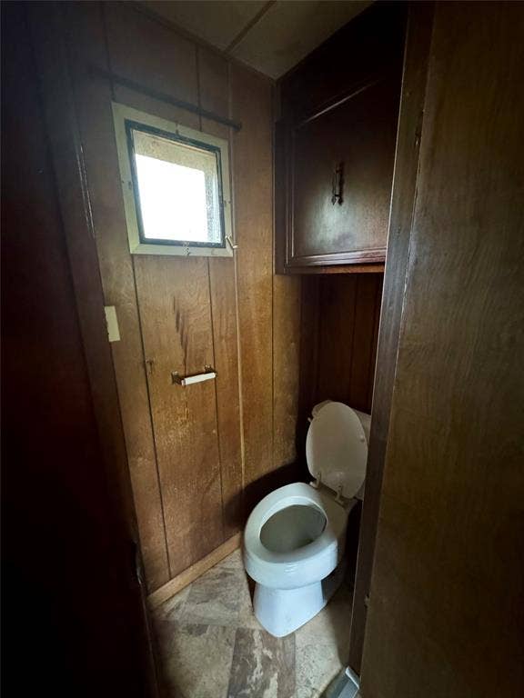 Wood-panelled walls, square window, built-in wooden upper cabinetry, porcelain toilet, and tile flooring