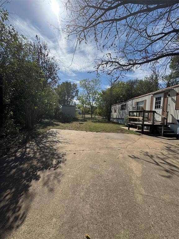 Concrete driveway, wood-frame deck, and expansive grass lot featuring mature trees and a detached storage shed