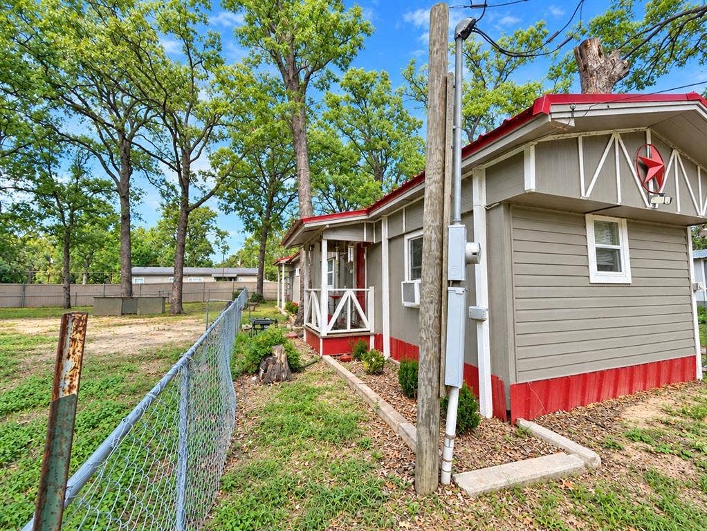 Exterior featuring horizontal siding, a gabled roofline with metal trim, a covered entryway with wood railing, and an expansive lot with mature trees and perimeter chain-link fencing