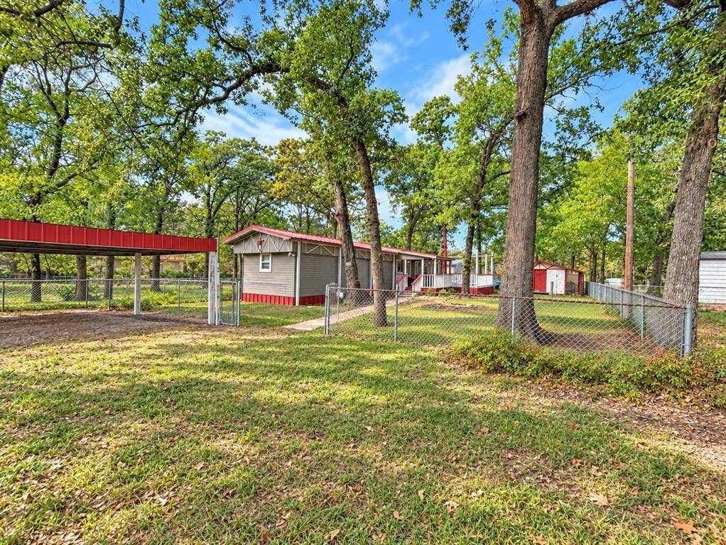 Detached carport with metal roofing, chain-link fencing, mature trees, and storage structure on a grass-covered lot