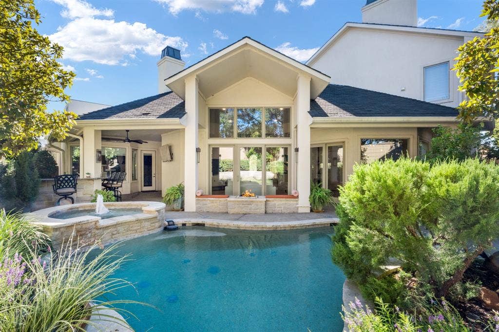 Stucco exterior featuring a gabled roofline, a built-in stone-clad fire feature, a covered patio with an integrated ceiling fan, and a custom swimming pool with an elevated stone spa