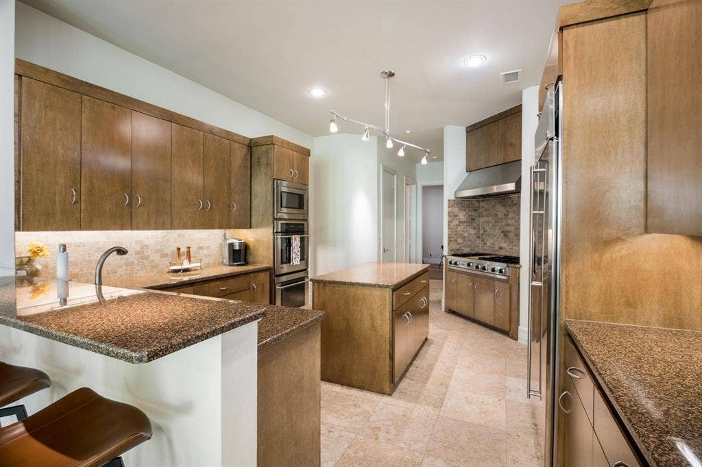 Kitchen featuring custom wood cabinetry, natural stone countertops, a multi-burner gas range with a stainless steel vent hood, stone tile backsplash, and tile flooring
