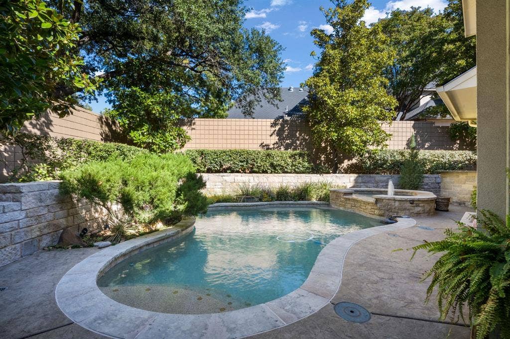 Curved swimming pool featuring a stone-finish spa, integrated water feature, textured concrete decking, and stacked stone retaining walls
