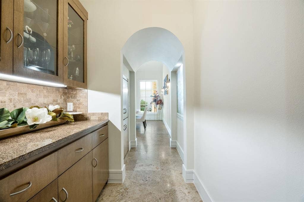 Built-in cabinetry with glass-front display uppers, stone-finish countertops, tiled backsplash, arched entryway, and natural stone flooring