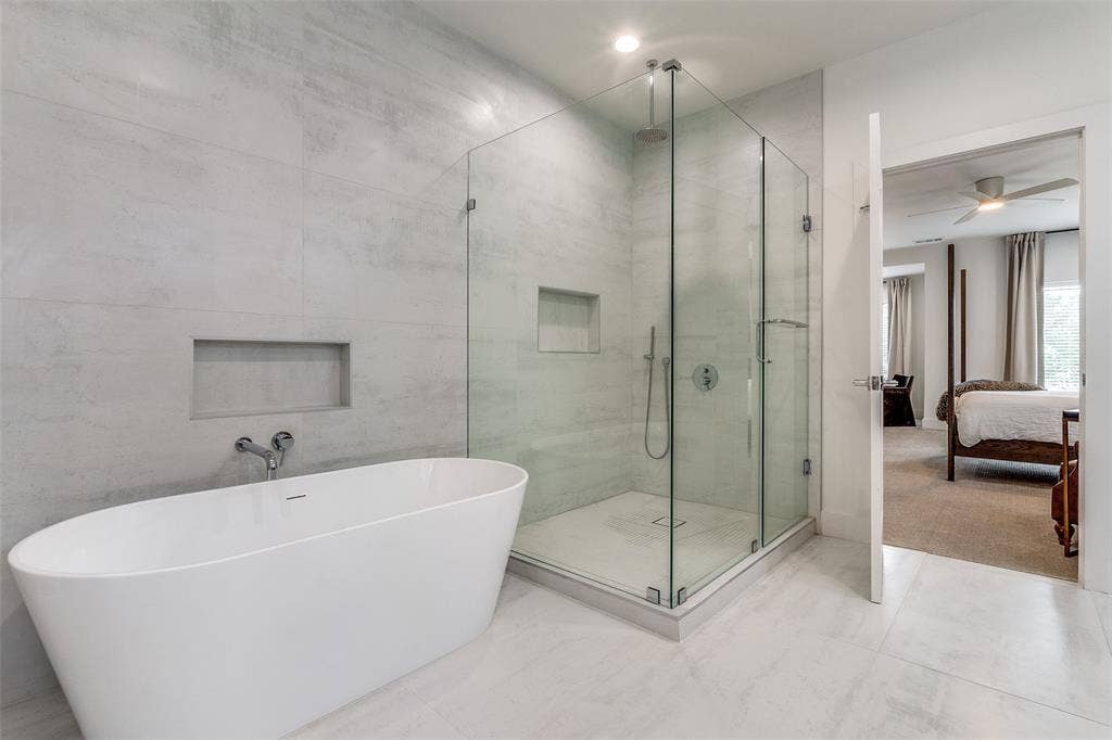 Freestanding white soaking tub with wall-mounted faucet, glass-enclosed walk-in shower with rainfall showerhead, and neutral large-format tile surround with recessed wall niches