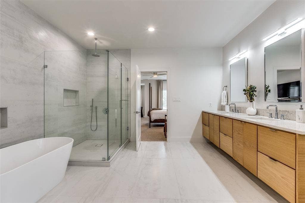 Dual-vanity cabinetry with light wood-finish, stone-top surface, glass-enclosed shower with tiled surround, and freestanding soaking tub