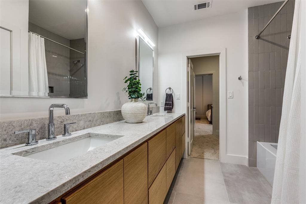 Bathroom featuring a long wood-finish vanity with dual undermount sinks, quartz countertop, polished chrome fixtures, and vertical tile shower surround