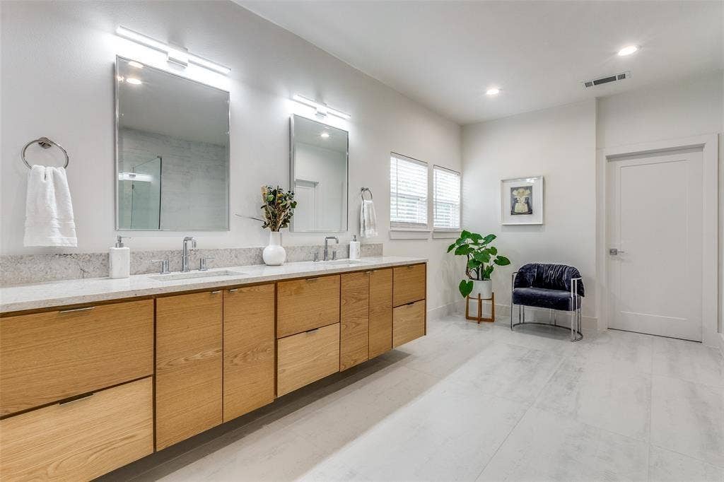 Dual-vanity bathroom featuring light-toned wood cabinetry, stone countertops, rectangular undermount sinks, integrated wall-mounted lighting, and large-format tile flooring
