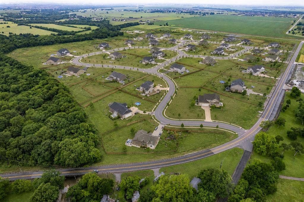 Aerial perspective of a residential community featuring winding paved roads, expansive open green space, mature wooded borders, and individual detached dwellings