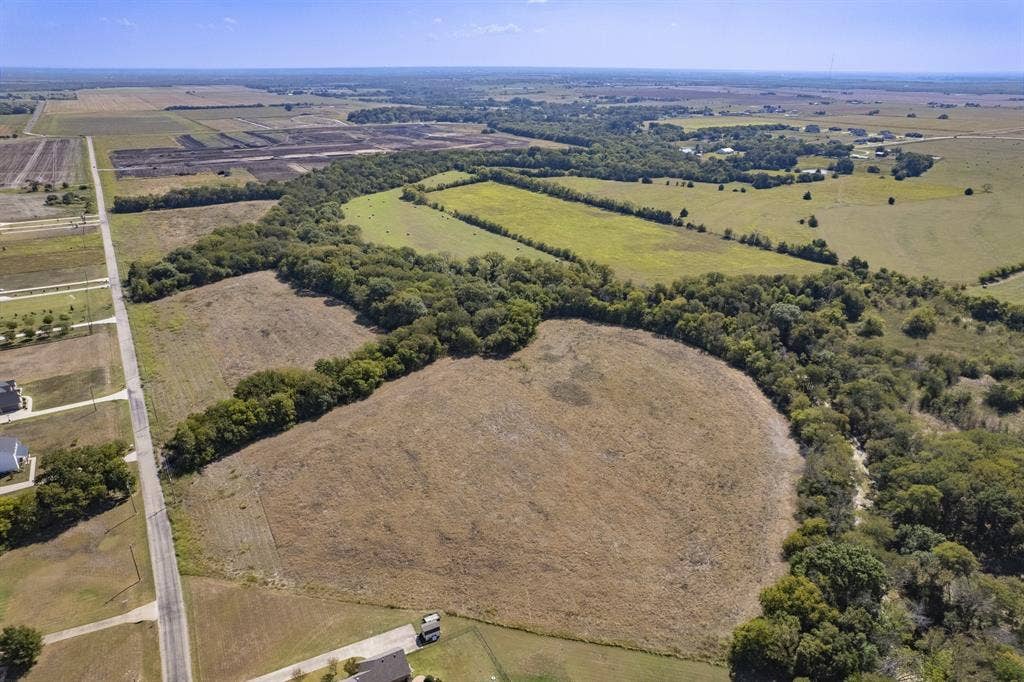 Aerial perspective of expansive acreage featuring open cleared fields, mature tree lines, and paved road access