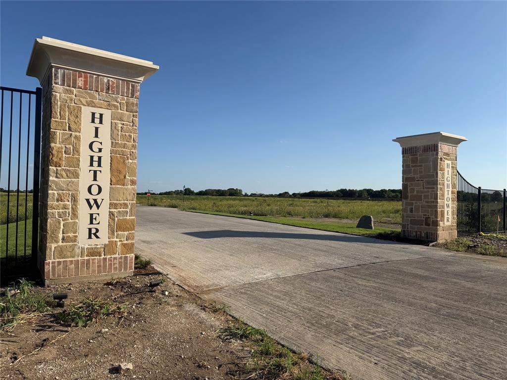 Gated entrance featuring custom masonry stone pillars with integrated cast stone signage, concrete driveway, and black metal perimeter fencing