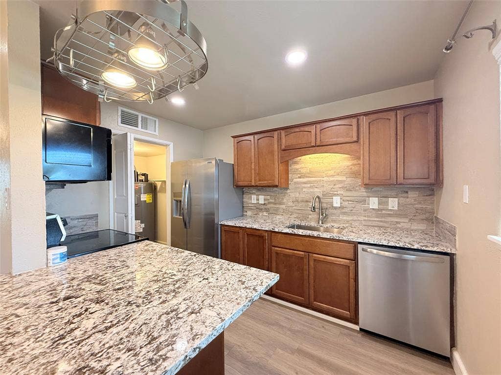 Kitchen featuring stained wood cabinetry, stone-patterned countertops, a stacked stone tile backsplash, stainless steel appliances, and a circular metal pot rack fixture