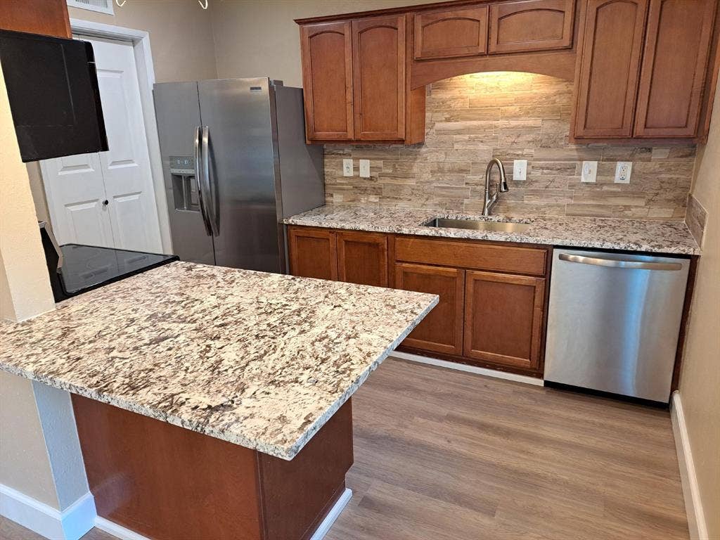 Kitchen featuring wood cabinetry, stone-finish countertops, stacked-stone backsplash, stainless steel appliances, and wood-finish flooring