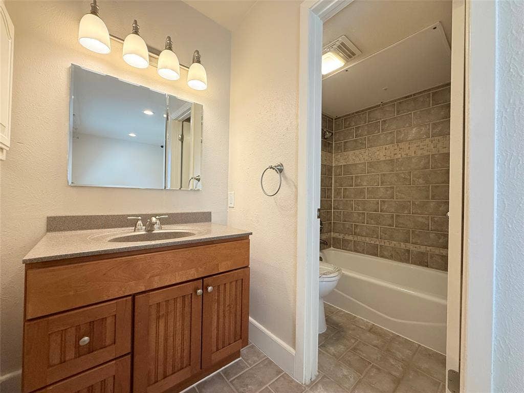 Bathroom vanity with wood-finish cabinetry, stone-composite countertop, wall-mounted four-light fixture, and tiled tub surround with integrated accent border