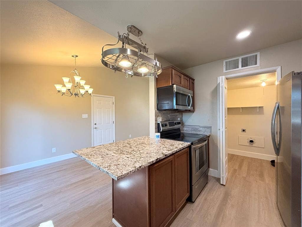 Kitchen featuring wood-finish flooring, stone-surface island, wood cabinetry, stainless steel appliances, and decorative overhead lighting fixtures