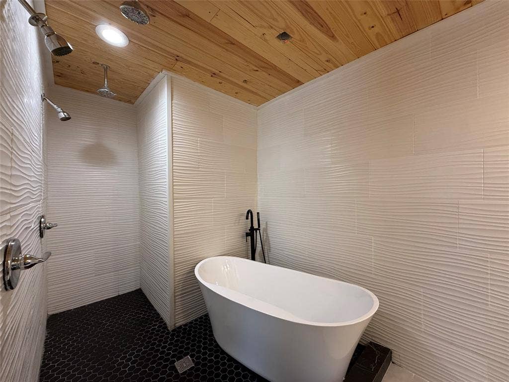2703 Downing Avenue, Dallas, TX 75216 Freestanding soaking tub with matte black fixtures, white textured wall tile, wood-plank ceiling, and black hexagonal floor tile, complemented by dual overhead shower heads