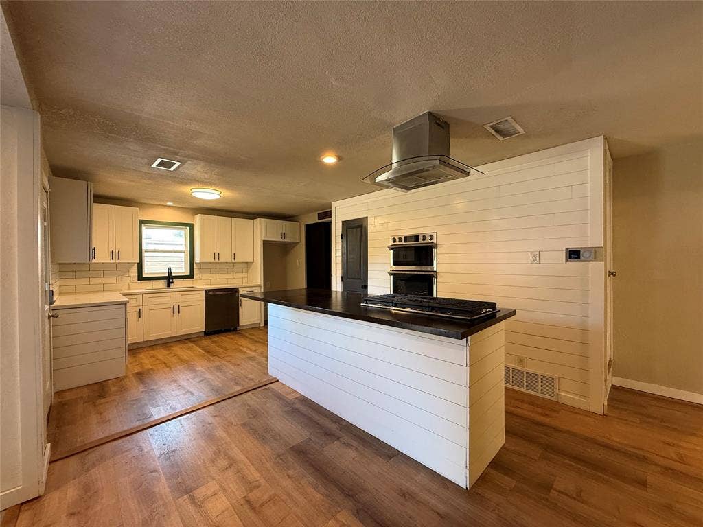 2703 Downing Avenue, Dallas, TX 75216 Kitchen featuring white shaker-style cabinetry, wood-finish flooring, a central island with a dark countertop and inset cooktop, a stainless steel range hood, and a built-in double wall oven