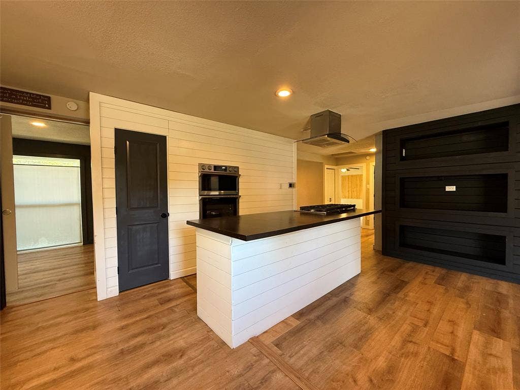 2703 Downing Avenue, Dallas, TX 75216 Kitchen featuring a central island with a dark countertop, stainless steel wall-mounted oven, stainless steel range hood, white horizontal shiplap wall paneling, and wood-finish flooring
