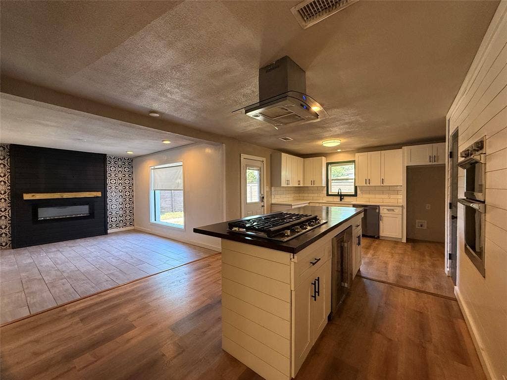 2703 Downing Avenue, Dallas, TX 75216 Kitchen featuring a shiplap-wrapped island with an integrated gas cooktop, built-in double wall oven, stainless steel range hood, and white cabinetry paired with a black fireplace accent wall