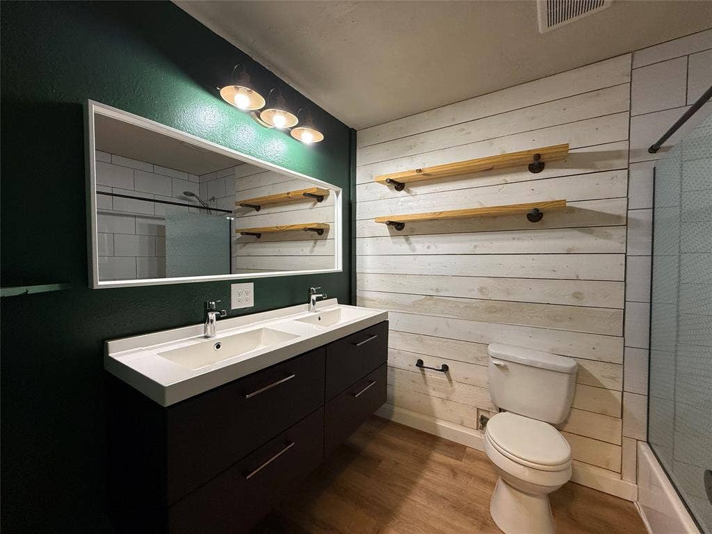 2703 Downing Avenue, Dallas, TX 75216 Dual-sink vanity with a rectangular countertop, white plank-style wall detailing, built-in wooden shelving, wood-finish flooring, and a large wall-mounted mirror